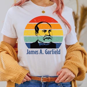 May include: White t-shirt featuring a retro design of James A. Garfield within a colorful circular graphic. The design includes a portrait of Garfield and his name in blue text. The shirt is a casual wear item.