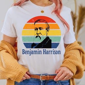 May include: White t-shirt featuring a retro design of Benjamin Harrison within a colorful sunset circle. The design includes a portrait and the name "Benjamin Harrison" in blue text. The shirt is a casual wear item.