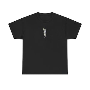 Könnte beinhalten: Ein schwarzes kurzärmeliges T-Shirt mit einer kleinen, mittig platzierten Grafik einer Figur in weißem Anzug, blauem Hemd und weißem Hut, die einen Arm hebt. Das Motiv befindet sich im oberen Brustbereich des einfarbigen schwarzen Stoffes.