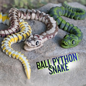 May include: Three articulated ball python snake toys in yellow, brown, and green, resting on a stone surface. The yellow snake has white accents, the brown snake has tan and white accents, and the green snake has dark green accents. The text "BALL PYTHON SNAKE" is visible.