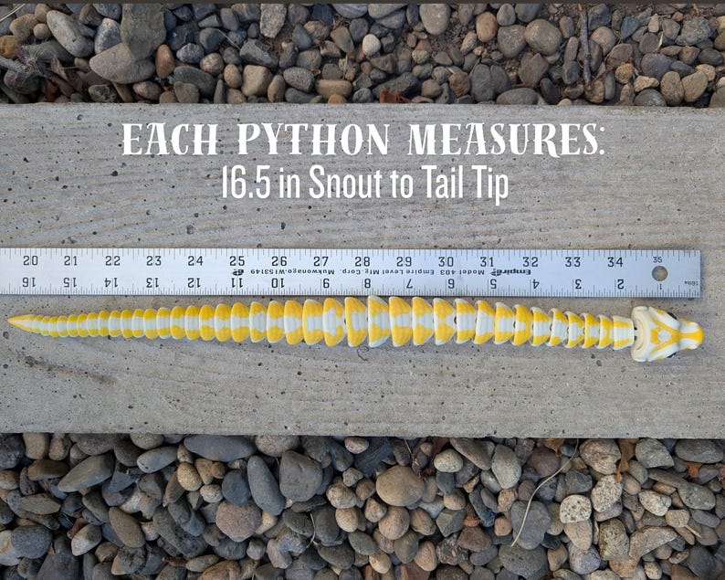 Pode incluir: Um modelo de p&iacute;ton amarelo e branco, com 42 cm de comprimento, &eacute; exibido em uma superf&iacute;cie de concreto ao lado de uma r&eacute;gua. O texto "EACH PYTHON MEASURES: 16.5 in Snout to Tail Tip" &eacute; vis&iacute;vel acima do modelo.
