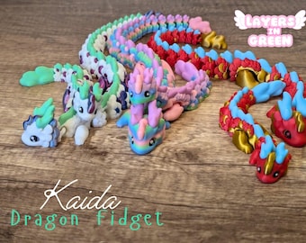Può includere: Un set di cinque giocattoli fidget a forma di drago colorati. Ogni drago è realizzato in plastica segmentata e flessibile e presenta uno schema di colori diverso. I draghi sono disposti in fila su una superficie di legno. Il testo "Kaida Dragon Fidget" è visibile nell'angolo inferiore sinistro dell'immagine. Il testo "Layers in Green" è visibile nell'angolo superiore destro dell'immagine.
