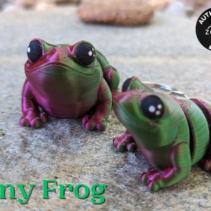May include: Two tiny green and purple frog figurines with black eyes. The frog on the left is sitting up, while the frog on the right is sitting with its head tilted up. The frogs are on a gray textured surface. The text "Tiny Frog" is printed in green below the frogs.