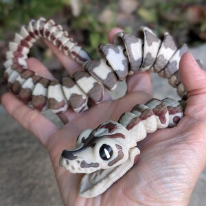 Pode incluir: Uma cobra de brinquedo articulada com um corpo creme, segmentos com padr&otilde;es castanhos e brancos e marcas castanho-avermelhadas. A cabe&ccedil;a da cobra &eacute; creme com marcas castanhas e boca aberta. O brinquedo &eacute; segurado em uma m&atilde;o.