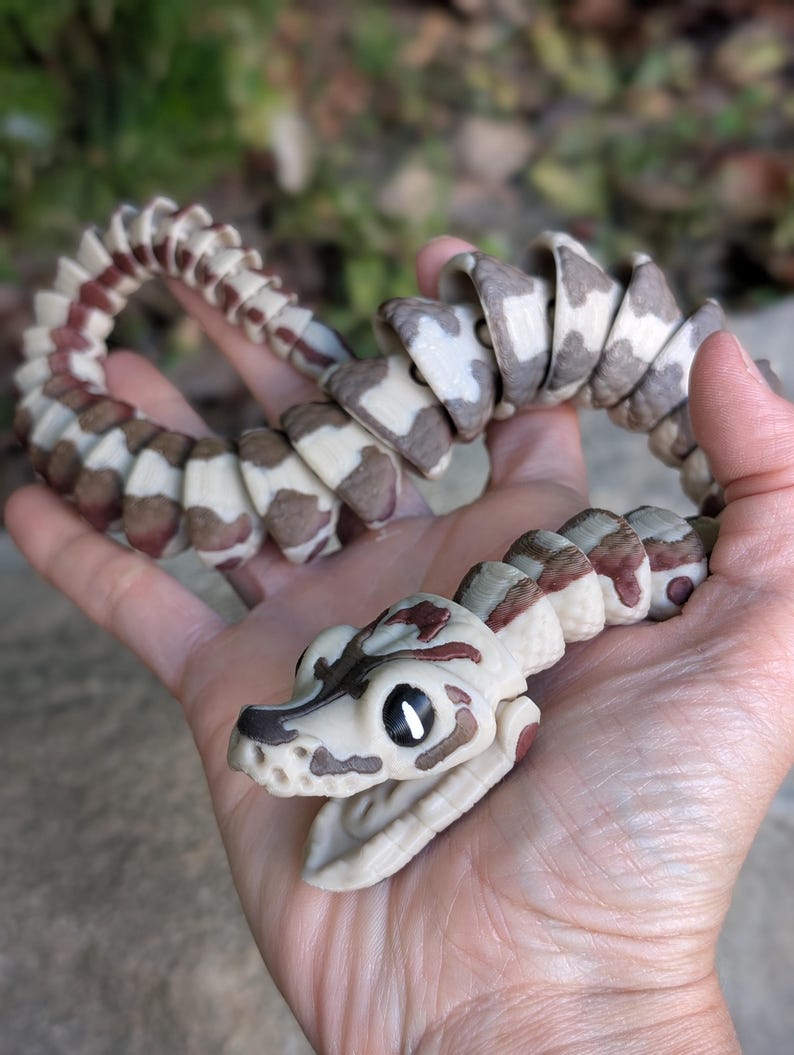 Pode incluir: Uma cobra de brinquedo articulada com um corpo creme, segmentos com padr&otilde;es castanhos e brancos e marcas castanho-avermelhadas. A cabe&ccedil;a da cobra &eacute; creme com marcas castanhas e boca aberta. O brinquedo &eacute; segurado em uma m&atilde;o.