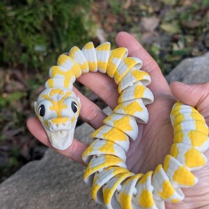 Pode incluir: Uma cobra de brinquedo segmentada em amarelo e branco, segurada em uma m&atilde;o. A cabe&ccedil;a da cobra tem olhos pretos e uma boca sorridente. Os segmentos do corpo alternam padr&otilde;es amarelos e brancos.