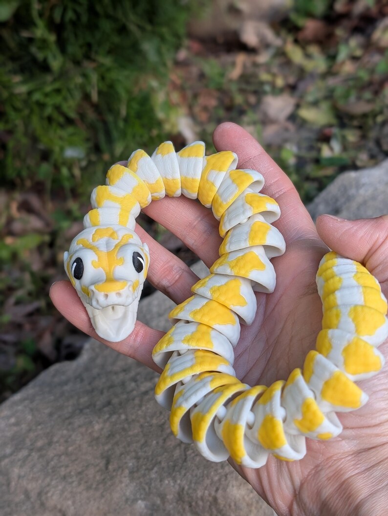 Pode incluir: Uma cobra de brinquedo segmentada em amarelo e branco, segurada em uma m&atilde;o. A cabe&ccedil;a da cobra tem olhos pretos e uma boca sorridente. Os segmentos do corpo alternam padr&otilde;es amarelos e brancos.