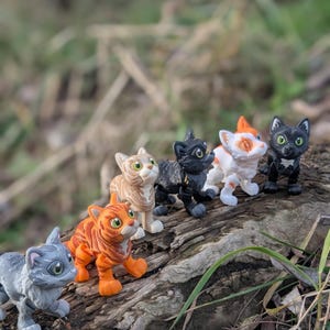 以下が含まれることがあります： 様々な色合いの6匹の小さな猫のフィギュアが、風化した丸太の上に並んでいます。グレー、オレンジ、黒など。それぞれの猫は詳細な特徴を持ち、遊び心のあるポーズをとっています。背景はぼやけており、屋外の環境を示唆しています。