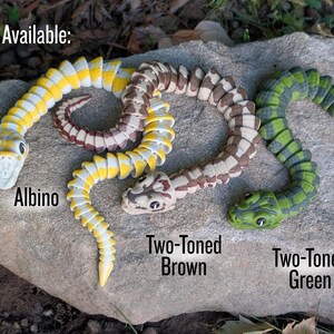 Pode incluir: S&atilde;o mostradas tr&ecirc;s cobras de brinquedo articuladas. Uma &eacute; amarela e branca, rotulada como "Albino". Outra &eacute; marrom e bege, rotulada como "Two-Toned Brown". A terceira &eacute; verde e preta, rotulada como "Two-Toned Green". A palavra "Available:" est&aacute; no topo.