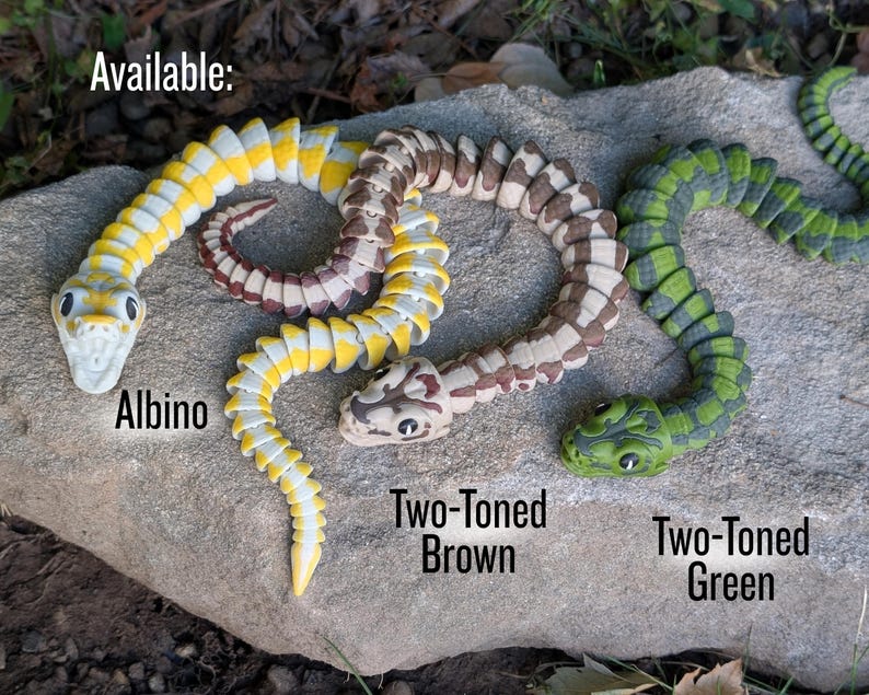 Pode incluir: S&atilde;o mostradas tr&ecirc;s cobras de brinquedo articuladas. Uma &eacute; amarela e branca, rotulada como "Albino". Outra &eacute; marrom e bege, rotulada como "Two-Toned Brown". A terceira &eacute; verde e preta, rotulada como "Two-Toned Green". A palavra "Available:" est&aacute; no topo.