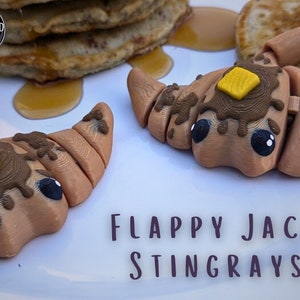 May include: Two brown and tan pancake stingray toys with yellow butter on top. The toys are on a white plate with the text "FLAPPY JACK STINGRAYS" below them.