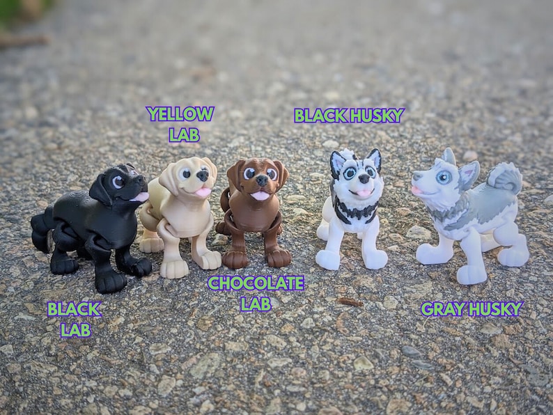Może przedstawiać: Pięć plastikowych figurek ps&oacute;w, w tym czarny labrador, ż&oacute;łty labrador, czekoladowy labrador, czarny husky i szary husky. Figurki stoją na szarej powierzchni.