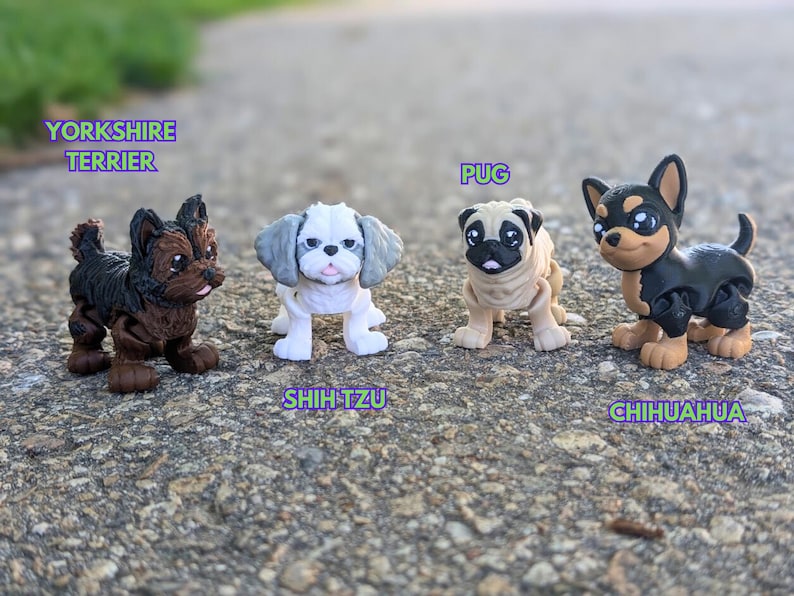 Może przedstawiać: Cztery małe plastikowe zabawki w kształcie ps&oacute;w, każda z innej rasy: Yorkshire Terrier, Shih Tzu, Mops i Chihuahua. Psy stoją na szarej i brązowej powierzchni.