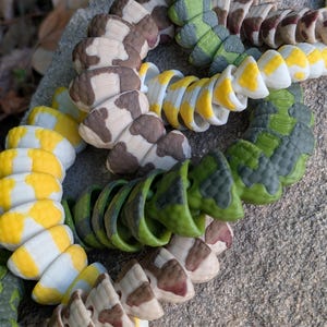 Pode incluir: Close-up de v&aacute;rios brinquedos coloridos em forma de cobra segmentada. Os brinquedos apresentam uma variedade de padr&otilde;es, incluindo manchas marrons e brancas, listras amarelas e brancas e camuflagem verde. Os brinquedos est&atilde;o dispostos em um padr&atilde;o circular.