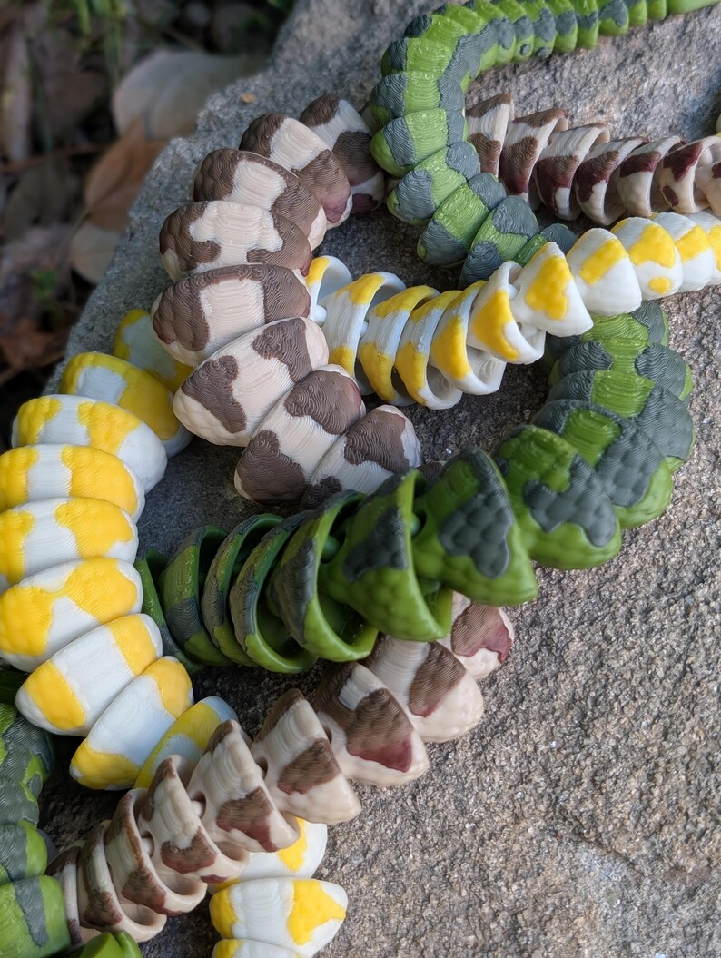 Pode incluir: Close-up de v&aacute;rios brinquedos coloridos em forma de cobra segmentada. Os brinquedos apresentam uma variedade de padr&otilde;es, incluindo manchas marrons e brancas, listras amarelas e brancas e camuflagem verde. Os brinquedos est&atilde;o dispostos em um padr&atilde;o circular.