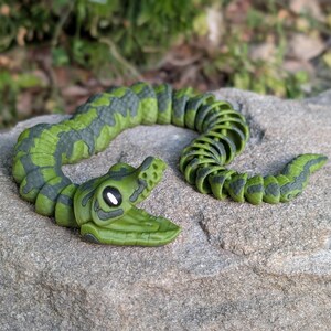 Pode incluir: Um brinquedo de cobra articulada verde e cinza. O brinquedo &eacute; feito de pl&aacute;stico e tem um corpo segmentado que lhe permite mover-se e dobrar-se. A cabe&ccedil;a da cobra &eacute; detalhada com olhos e boca. A cobra est&aacute; pousada numa superf&iacute;cie de pedra cinzenta.