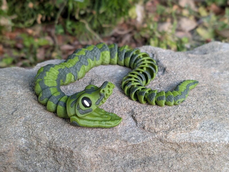 Pode incluir: Um brinquedo de cobra articulada verde e cinza. O brinquedo &eacute; feito de pl&aacute;stico e tem um corpo segmentado que lhe permite mover-se e dobrar-se. A cabe&ccedil;a da cobra &eacute; detalhada com olhos e boca. A cobra est&aacute; pousada numa superf&iacute;cie de pedra cinzenta.