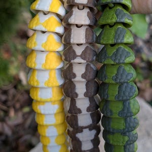 Pode incluir: Tr&ecirc;s fios de pe&ccedil;as decorativas segmentadas em amarelo e branco, castanho e branco e padr&otilde;es de camuflagem verde e cinzento. As pe&ccedil;as est&atilde;o ligadas, assemelhando-se &agrave; cauda de uma cobra ou a uma corrente decorativa.