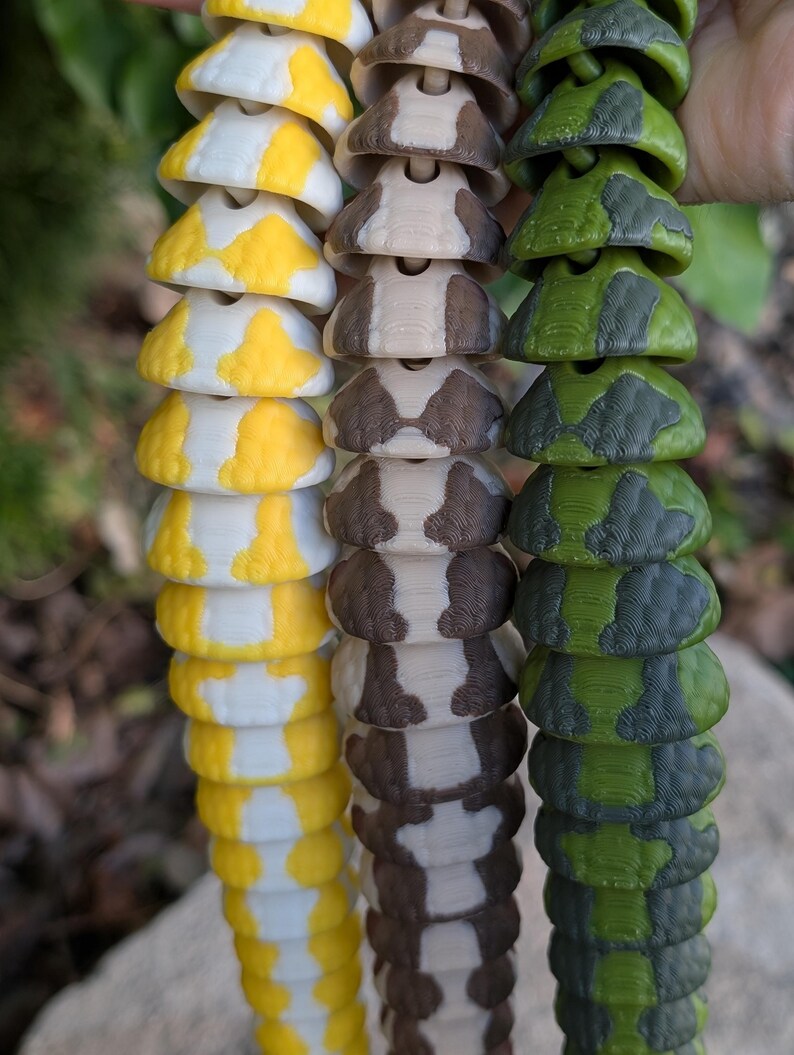 Pode incluir: Tr&ecirc;s fios de pe&ccedil;as decorativas segmentadas em amarelo e branco, castanho e branco e padr&otilde;es de camuflagem verde e cinzento. As pe&ccedil;as est&atilde;o ligadas, assemelhando-se &agrave; cauda de uma cobra ou a uma corrente decorativa.