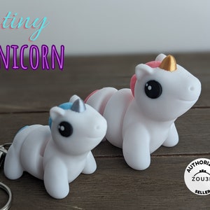 May include: Two white unicorn keychains with gold horns. One unicorn has a pink mane and the other has a blue mane. The keychains are on a wooden surface. The text "tiny UNICORN" is in the upper left corner of the image. The text "AUTHORIZED ZOU3D SELLER" is in a circle in the lower right corner of the image.