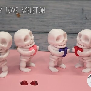 May include: Four white skeleton figurines holding different colored hearts. The skeletons are small and have a cute, cartoonish appearance. The hearts are pink, purple, red, and black. The background is a light pink surface. The text "TINY LOVE SKELETON" is at the top of the image.