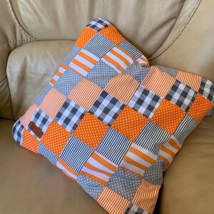 May include: A patchwork cushion with orange and grey squares. The cushion is made of different fabrics with various patterns, including stripes, polka dots and plaid.