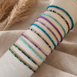 Op de afbeelding: Een collectie van tien dunne, delicate armbanden met kleine, gefacetteerde, veelkleurige edelsteenkralen. De armbanden zijn in een stapel gerangschikt en tonen een verscheidenheid aan tinten, waaronder helderblauw, paars, turkoois en aardse gespikkelde patronen.