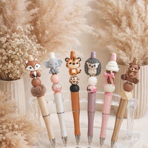 Peut inclure: Ensemble de six stylos décoratifs avec des motifs d'animaux et de desserts. Les stylos présentent diverses couleurs, notamment beige, blanc, orange, violet et rose. Chaque stylo mesure environ 15 cm de long et est présenté dans un support en acrylique transparent.