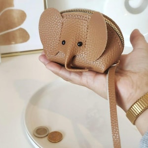 Peut inclure: Un porte-monnaie marron en forme d'éléphant avec une fermeture éclair et une petite sangle. Le porte-monnaie a des yeux noirs et de grandes oreilles. Le porte-monnaie est fait d'un matériau texturé.