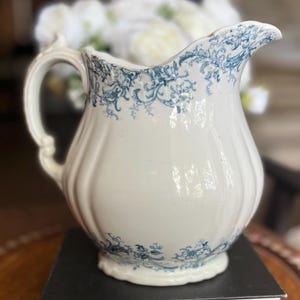 May include: A cream-colored ceramic pitcher featuring a ribbed body and a decorative blue floral pattern along the rim and base. The pitcher has a curved handle and a wide pouring spout, with a smooth, glossy finish throughout the exterior surface.