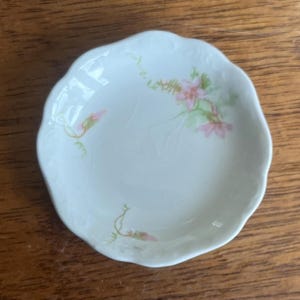 May include: A small, white, scalloped-edge dish with a floral design. The dish features pink and green flowers and buds, with delicate green stems and leaves. The dish is likely made of porcelain or ceramic.