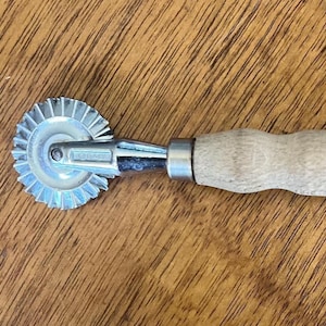 May include: A sewing tracing wheel featuring a serrated metal wheel with a silver finish, a chrome-plated metal shank, and a light-colored wooden handle with a contoured grip. The metal shank is clearly stamped with the word ITALY.