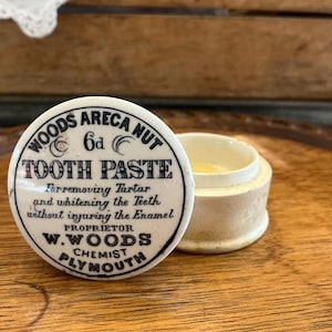 May include: Vintage Woods Areca Nut Tooth Paste jar with lid. The white lid has black text that reads "Tooth Paste" and "Plymouth Chemist". The jar is open, revealing a yellow paste inside. The jar sits on a wooden surface.
