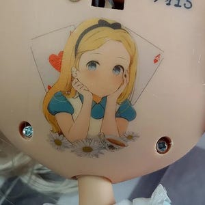May include: A light beige circular object featuring a printed illustration of a blonde character with a black headband, wearing a blue dress, resting their chin on their hands. The background includes a red heart playing card and white daisies.