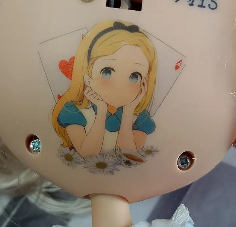 May include: A light beige circular object featuring a printed illustration of a blonde character with a black headband, wearing a blue dress, resting their chin on their hands. The background includes a red heart playing card and white daisies.