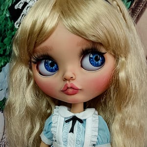 May include: A doll with long blonde hair, blue eyes, and long eyelashes, wearing a black headband with white lace trim. The doll is dressed in a light blue dress with a white pinafore featuring a small black bow at the centre of the neckline.