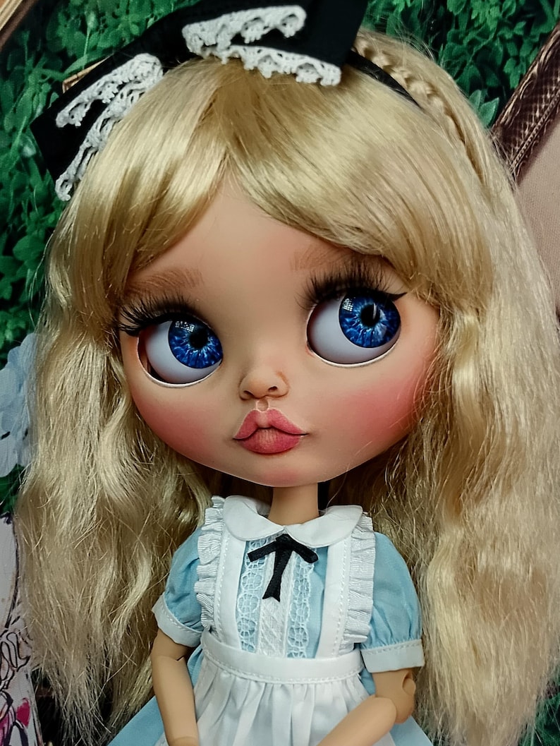 May include: A doll with long blonde hair, blue eyes, and long eyelashes, wearing a black headband with white lace trim. The doll is dressed in a light blue dress with a white pinafore featuring a small black bow at the centre of the neckline.