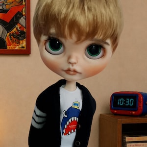 Może przedstawiać: Lalka z kr&oacute;tkimi blond włosami i dużymi zielonymi oczami, ubrana w biały t-shirt z grafiką niebieskiego rekina z małą ż&oacute;łtą postacią na g&oacute;rze, pod czarnym rozpinanym kardiganem z szarymi paskami na rękawach oraz niebieskimi dżinsami.