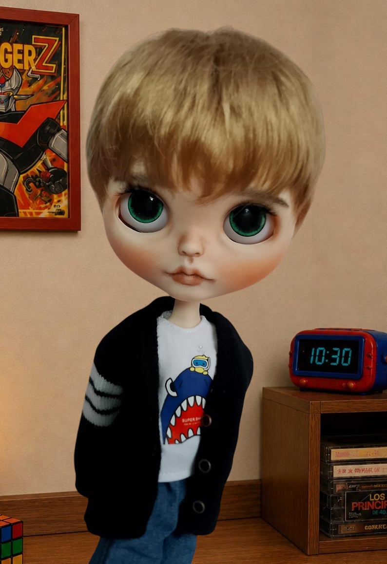 Może przedstawiać: Lalka z kr&oacute;tkimi blond włosami i dużymi zielonymi oczami, ubrana w biały t-shirt z grafiką niebieskiego rekina z małą ż&oacute;łtą postacią na g&oacute;rze, pod czarnym rozpinanym kardiganem z szarymi paskami na rękawach oraz niebieskimi dżinsami.