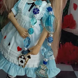 May include: A doll wearing a light blue dress with white lace trim and a black bow, adorned with a long beaded necklace featuring blue and white beads, a spade-shaped playing card charm, and a miniature dress-shaped metal pendant.