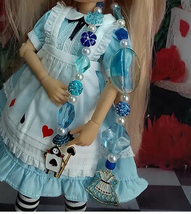 May include: A doll wearing a light blue dress with white lace trim and a black bow, adorned with a long beaded necklace featuring blue and white beads, a spade-shaped playing card charm, and a miniature dress-shaped metal pendant.