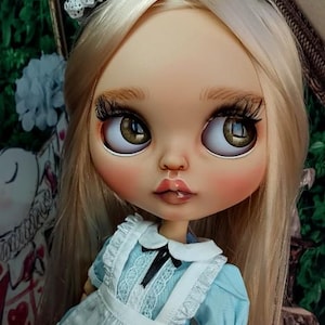 May include: A doll with long blonde hair, brown eyes, and long eyelashes, wearing a light blue dress with a white apron, a white collar, and a black bow. The doll has a black headband with white lace trim on its head and a neutral facial expression.