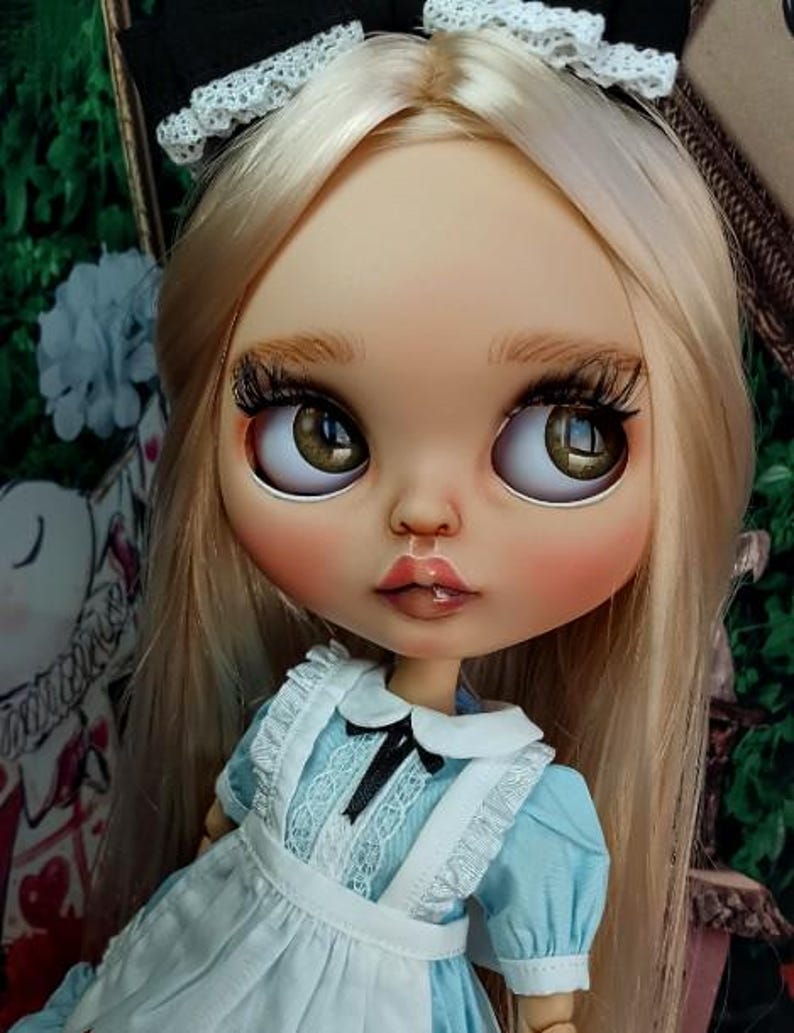 May include: A doll with long blonde hair, brown eyes, and long eyelashes, wearing a light blue dress with a white apron, a white collar, and a black bow. The doll has a black headband with white lace trim on its head and a neutral facial expression.