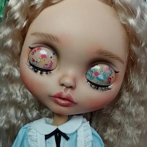 May include: A doll with long wavy blonde hair and large eyes featuring printed graphics of Alice in Wonderland characters and floral patterns. The doll wears a light blue dress with a white collar, white ruffled trim, and a black ribbon bow tie.