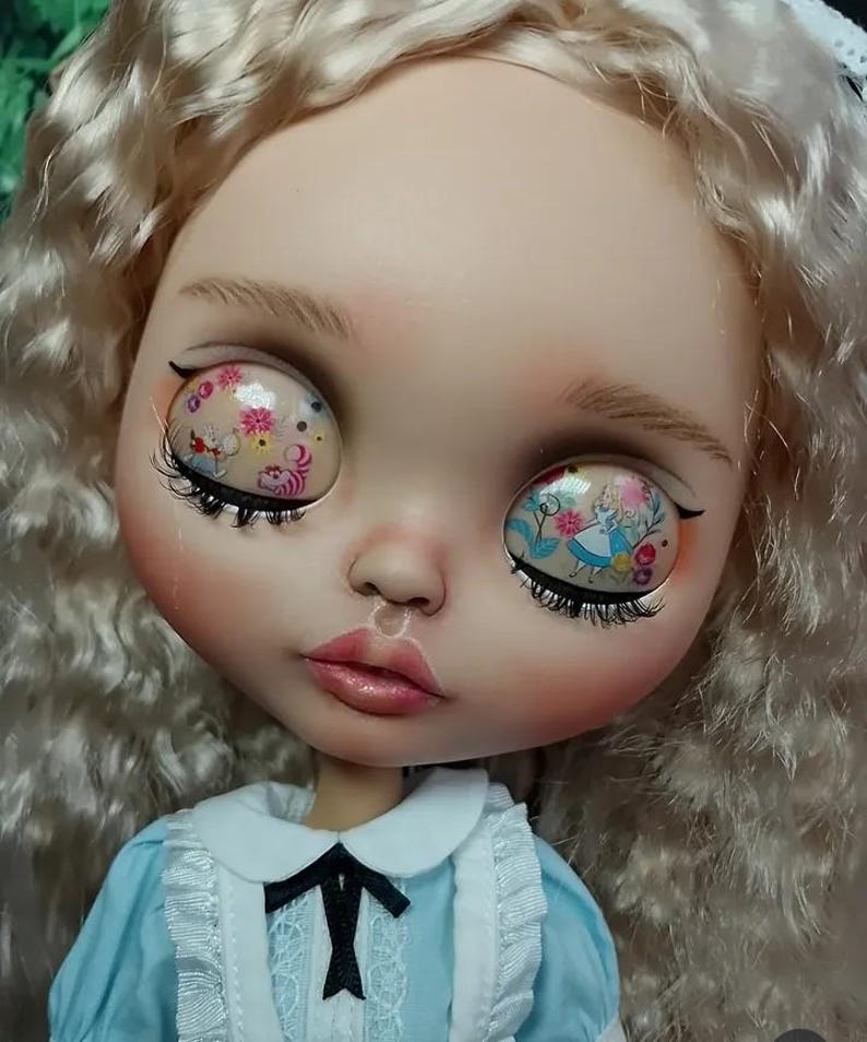 May include: A doll with long wavy blonde hair and large eyes featuring printed graphics of Alice in Wonderland characters and floral patterns. The doll wears a light blue dress with a white collar, white ruffled trim, and a black ribbon bow tie.