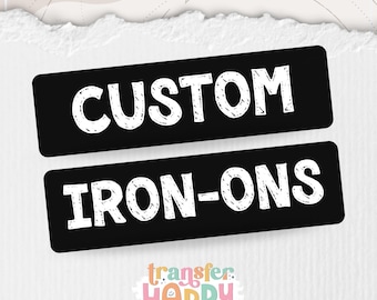 May include: Two rectangular black labels with white text that reads CUSTOM and IRON-ONS in a hand-drawn style font. Below the labels is a colourful logo with the text transfer HAPPY in a mix of orange, blue, yellow, and pink letters.