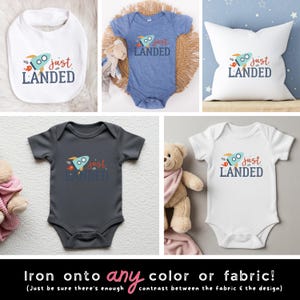 May include: A collection of baby apparel and a pillow featuring a graphic of a light blue rocket with the text "just LANDED" in red and dark blue. The items include a white bib, a heather blue bodysuit, a white pillow, a dark gray bodysuit, and a white bodysuit.