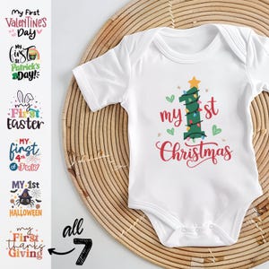 May include: A white short-sleeved baby bodysuit featuring a graphic of a green number one shaped like a Christmas tree with a yellow star, colorful lights, and the red text My 1st Christmas. Six additional holiday designs are shown on the left.