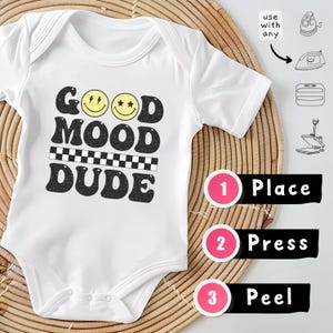 May include: A white baby bodysuit featuring a black graphic with the text GOOD MOOD DUDE, two yellow smiley faces, and a checkered line. Instructions include icons for an iron and heat press, with numbered steps: 1 Place, 2 Press, 3 Peel.
