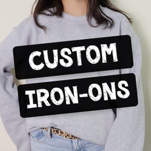 May include: A light gray crewneck sweatshirt featuring two horizontal black rectangular patches with white distressed-style text that reads CUSTOM on the top patch and IRON-ONS on the bottom patch, worn over light blue denim jeans with a leopard print belt.